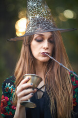 Young witch holding a wand and a goblet, preparing for a ritual, sitting alone in an autumn forest