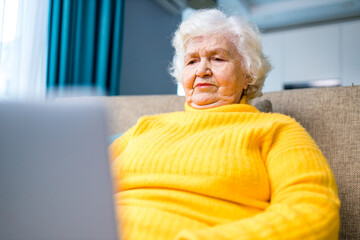 Happy mature blonde lady sitting on couch with laptop in living room