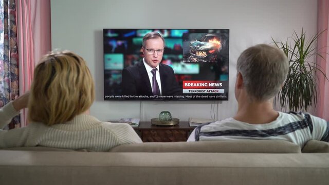 Family Couple Watching TV News Sitting on Couch in Living Room Together. TV Presenter Telling Breaking News about Terrorist Attack with Scenes Violence and Fire. Rear View