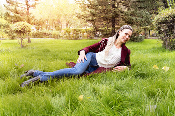 A young woman in a burgundy coat lies on the green grass and smiles. Wallow in the grass and enjoy life. Autumn mood. Freedom.

