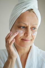 Close-up of a mature woman in a white bathrobe with a towel on her head applying cosmetic cream to her face