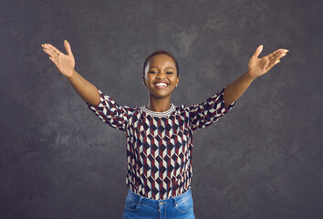 Happy cheerful black woman with short hair in trendy geometric pattern casual sweater spreads arms wide open to hug you, give someone warm welcome or present something standing against grey background