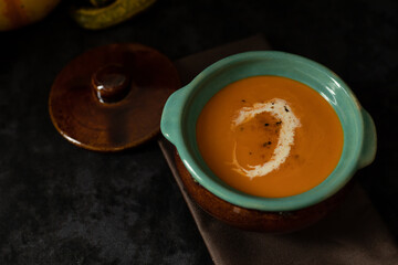 Pumpkin seasonal cream soup in a clay pot on the table, dark background with copy space