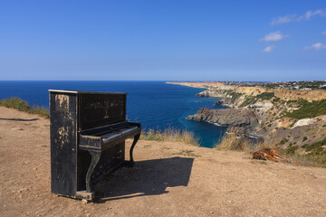 An old black piano and a dog lying next to him on a high rocky coast in the Crimea at Cape Fiolent. In the background there is a picturesque bay with blue water and blue sky. The city in the distance 