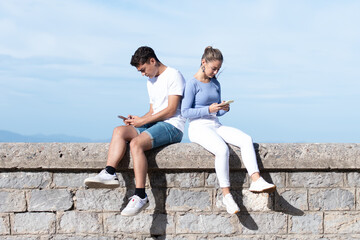 Fototapeta na wymiar Sad teenager couple concentrated on their phones sitting on a stone wall. Digital addiction and dopamine concept.