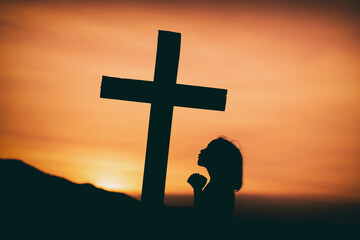 young girl standing praying under the cross with light of sunset background, christian silhouette...