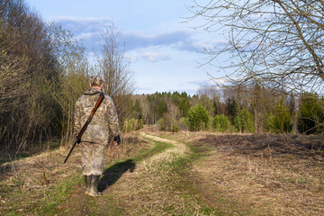 a hunter with a shotgun on his back walks along a forest road in spring