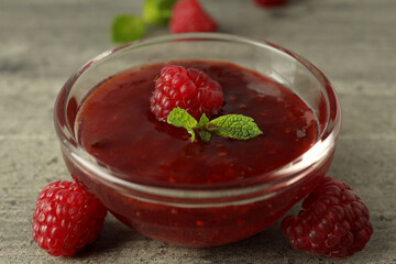 Glass bowl of raspberry jam with ingredients on gray textured background