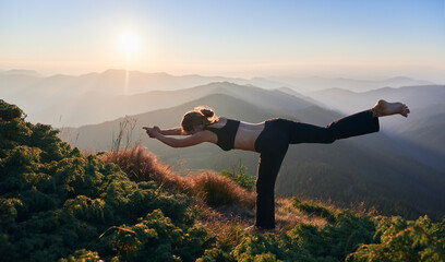 Side view of woman practicing yoga in fly pose outdoor. Meditating female is balancing on one leg in mountains after sunset. Concept of nature.