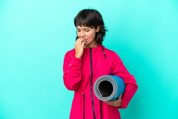 Fototapeta premium Young girl going to yoga classes while holding a mat isolated on blue background having doubts