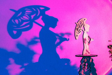 Tender small statue of young woman holding sun umbrella casting dark shadow on purple surface illuminated by bright spotlight beam