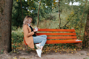 Side view of beautiful young blonde woman in coat having rest on hanging swing bench in nature...