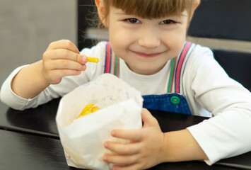 A cheerful, funny little girl is eating French fries while sitting at a table in a street fast food cafe. A happy child holds a potato in his hand.Unhealthy American food is popular all over the world