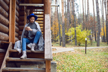 woman in a blue hat and scarf and a mug stands at a wooden house in the woods