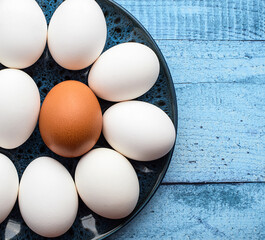 White raw eggs and one brown egg in a bowl on a blue wooden table, close up. Top view, flat lay. Farm products concept. Healthy breakfast.