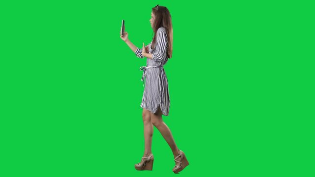 Side view of walking young woman greeting and waving during smart phone video call. Full body on green screen chroma key background