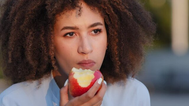 Extreme close-up portrait outdoors african american woman with curly hair afro hairstyle pensive lady female eating biting red fresh delicious natural apple enjoying eat fruit sad food diet chewing