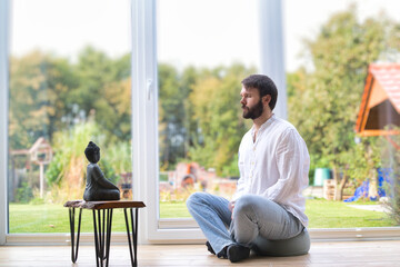 A bearded man is meditating cross legged sitting on a pillow in front of a buddha statue. He is wearing white shirt and has his eyes closed.