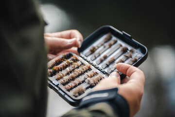Fly fisherman chooses artificial flies from lures box for fly fishing at river. Blurry close up. - Powered by Adobe