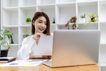 Beautiful Asian businesswoman sitting in her private office, chatting with her partner via laptop and checking documents, she is a female executive of a startup company. Financial management concept.