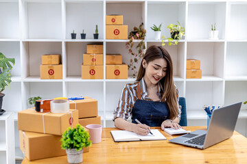 A beautiful Asian business owner opens an online store, she is checking orders from customers via laptop, sending goods through a courier company, concept of a woman opening an online business.