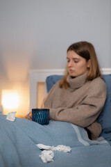 Exhausted caucasian woman in beige sweater sitting in bed and drinks warm medicine for sore throat. Girl suffering from cold, flu or angina. Selective focus on cup. Healthcare concept.