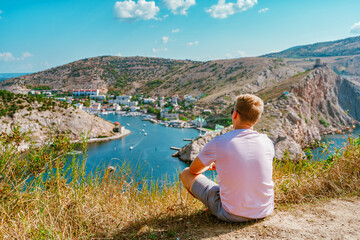 Fototapeta na wymiar A young blond man enjoys a picturesque landscape with a view of Balaclava with yachts and a colorful bay in summer. Postcard view of the tourist Crimea.