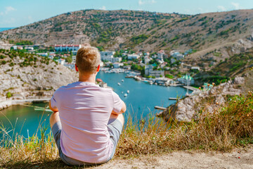 A young blond man enjoys a picturesque landscape with a view of Balaclava with yachts and a colorful bay in summer. Postcard view of the tourist Crimea.