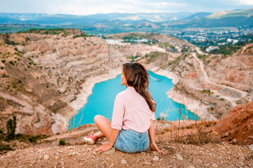 Naklejka na ściany i meble A young beautiful woman is sitting on the edge of a cliff with a Kadykovsky quarry in the shape of a heart in the Crimea. The concept of traveling in Russia