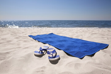 Flip flops and blue beach towel on sandy seashore - obrazy, fototapety, plakaty