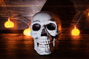 Human skull in front of luminous garland with halloween pumpkin lamps on dark wooden surface