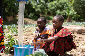 Two little black African girls in brightly colored clothes sitting on an arid cabbage field and patiently watching a broad jet of water that is about to fill their bucket
