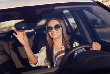 Plakat Photo of adorable cute young woman wear white shirt dark glasses riding car looking back mirror smiling outside city street