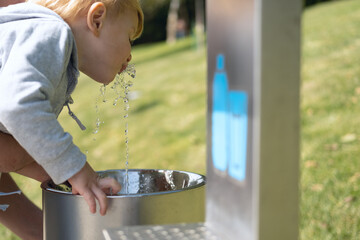 father helps his son kid boy to drink water from public drinking fountain in a park