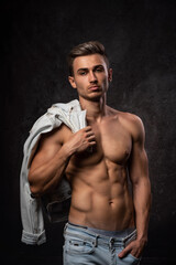 Fototapeta na wymiar European athletic young man, posing without outerwear, in sports physical form. On a dark textured background. Jacket on the shoulder.
