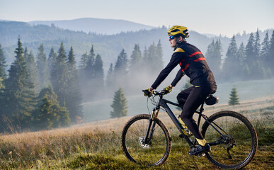 Fototapeta na wymiar Man bicyclist riding his bicycle in the mountains in early foggy morning. Side view of cyclist riding down a hill against beautiful landscape. Copy space. Concept of extreme sport