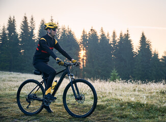 Fototapeta premium Side view of cyclist standing with bike on mountain trail with coniferous trees on background. Man bicyclist enjoying bicycle ride in mountains in the morning. Concept of sport and active leisure.