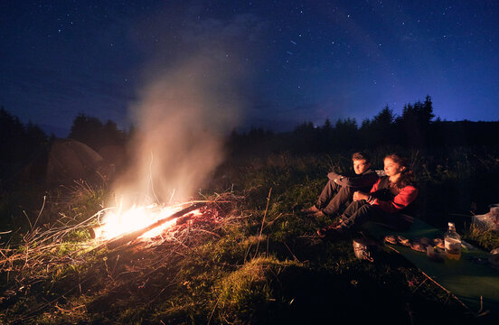 Horizontal Snapshot Of Couple Of Tourists Spending Time Together In Camping. Young Boy And Girl Near Campfire In The Evening Outdoors In Nature.