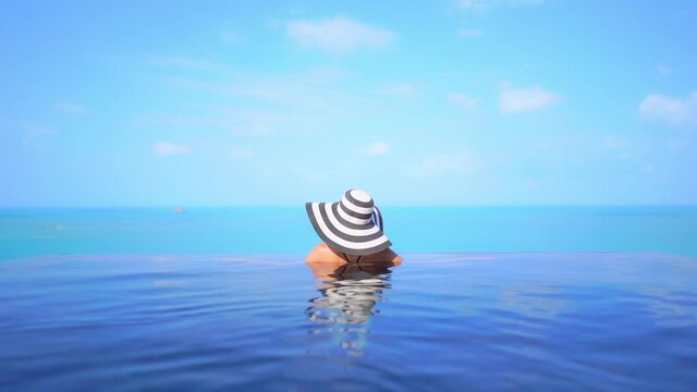 A woman in a floppy sun hat with her back to the camera looks out from her resort swimming pool at the ocean beyond. Title space