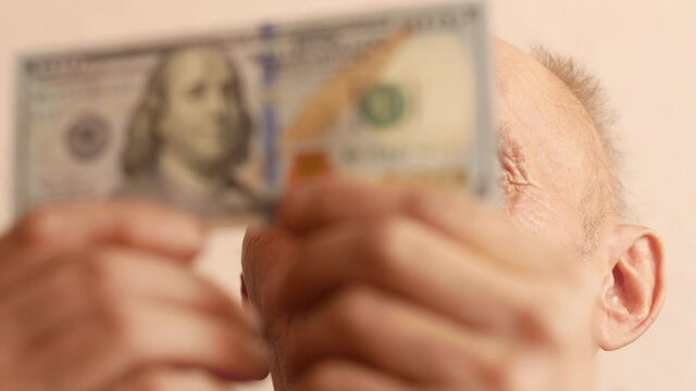 Portrait of a Caucasian pensioner 70 years older looking at a 100 dollar bill. Household budget and money of the elderly. Receiving dividends from a deposit.Selective focus, shallow depth of field