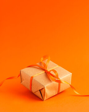Close up of brown gift box with a orange satin ribbon bow on orange blurred background with copy space. Holiday autumn concept .