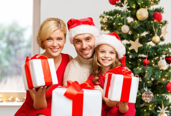 family, winter holidays and people concept - smiling mother, father and daughter in santa hats with gifts at home over christmas tree on background