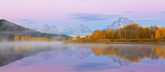 Fototapeta na wymiar Dawn image of the snowcapped Grand Tetons with mist, orange fall colors and perfect reflections in the river.