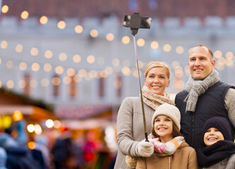 winter holidays, technology and people concept - happy family taking picture with smartphone and selfie stick over christmas market background