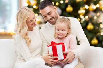 Fototapeta na wymiar family, winter holidays and people concept - happy mother, father and little daughter with gift box sitting on sofa at home over christmas tree background