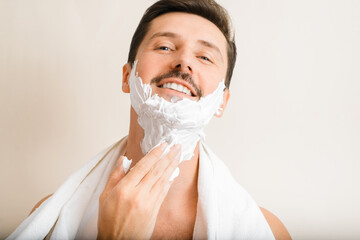 Close-up portrait of handsome positive man with towel on shoulders applying moisturizing shaving...