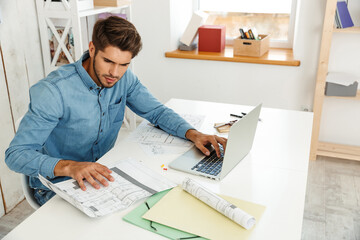 Young white man working with laptop and drawings in office