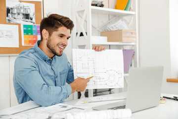 Fototapeta na wymiar Young white man smiling while working with laptop and drawings in office