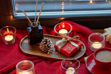 holidays, decoration and celebration concept - christmas gift, cup of coffee, candles and aroma reed diffuser on red tablecloth on window sill at home