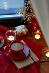 Obraz na płótnie Canvas holidays, decoration and celebration concept - cup of coffee in red ceramic cup with saucer, burning candles and christmas gift on window sill at home